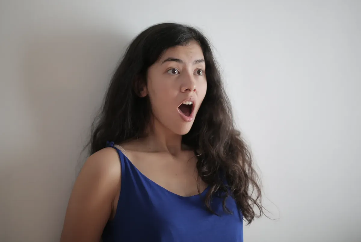 A surprised young woman with long brunette hair and an expressive face in a blue top.